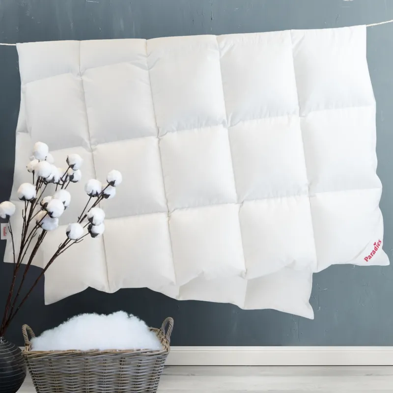 Weiße Steppdecke mit quadratischer Steppung an einer grauen Wand hängend, davor ein Korb mit Füllmaterial und ein Zweig mit Baumwollblüten