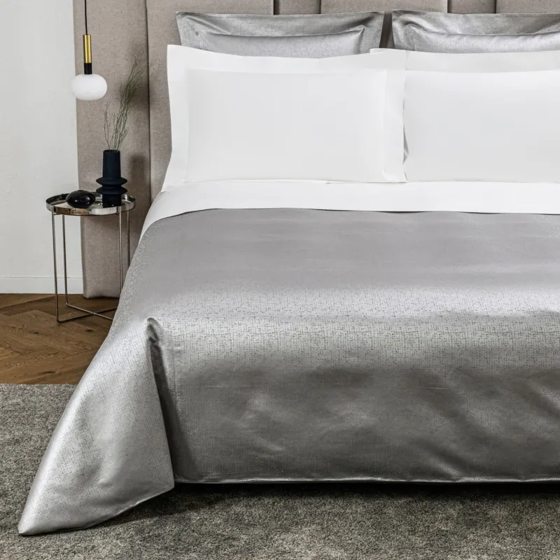 A neatly made bed features Frette’s Luxe Glowing Weave Duvet Cover, paired with white cotton sateen sheets and pillowcases. A small side table displays décor, while a pendant light hangs nearby, completing the serene setting.