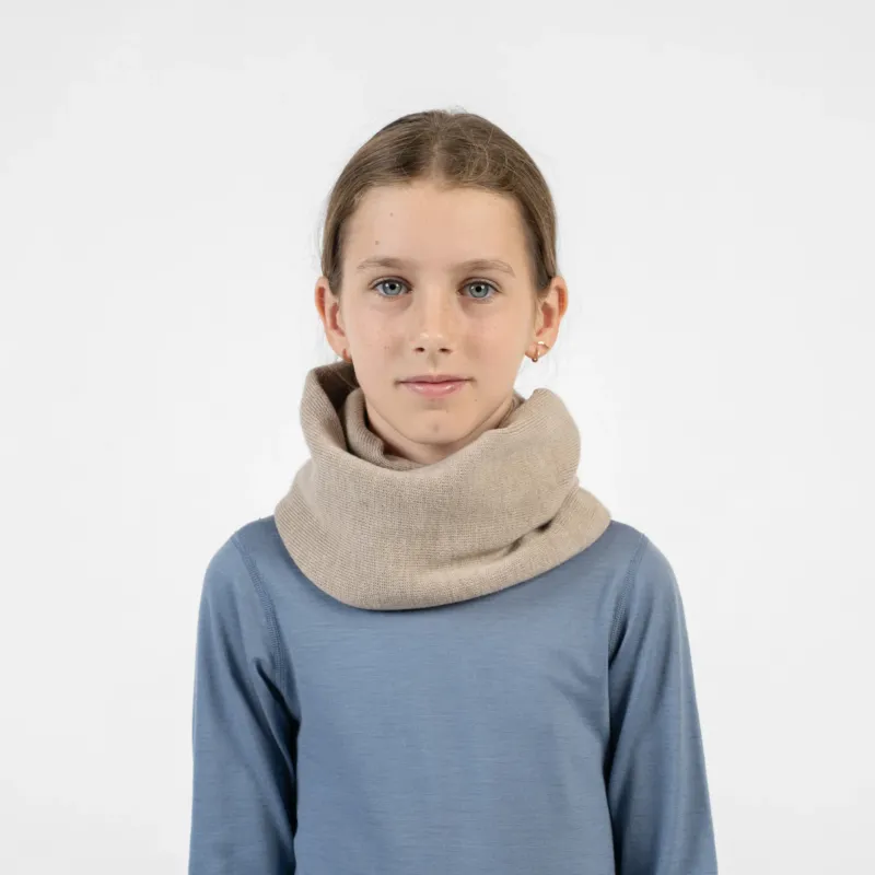 A young girl looking directly forward, wearing a beige knit infinity scarf around her neck and a blue long-sleeved shirt.