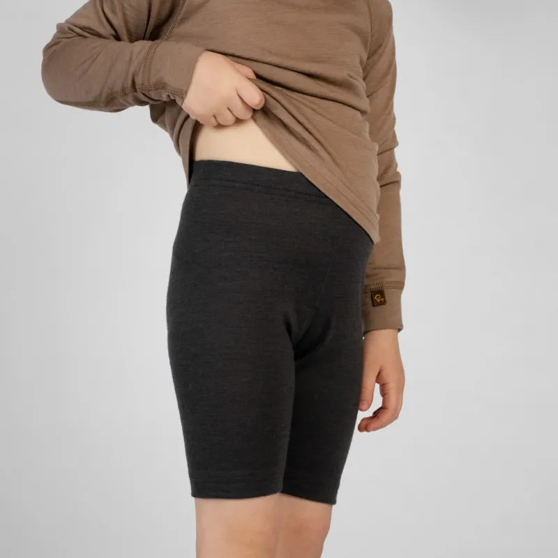 A slightly angled view of a child wearing dark charcoal merino wool cycling shorts paired with a brown long-sleeve top. The child is lifting the front of the shirt with one hand to display the high-waisted fit of the shorts, set against a plain light gray studio background.