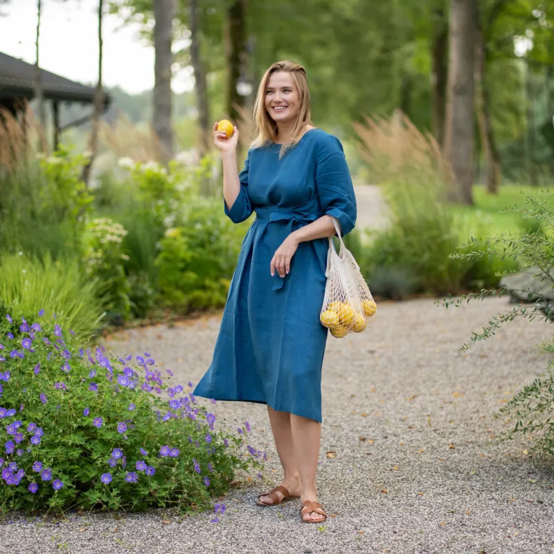 A smiling woman stands on a gravel garden path wearing a cobalt blue, three-quarter sleeve linen dress with a tied waist. She playfully holds up a single lemon in one hand while carrying a string bag full of lemons in the other.