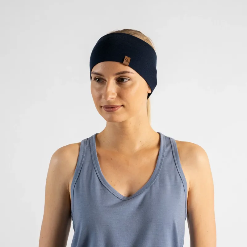 A studio portrait of a blonde woman looking down and off to the left side of the frame against a plain light grey background. She is wearing a dusty blue v-neck sleeveless top and a wide navy blue headband with a small brown logo tag.