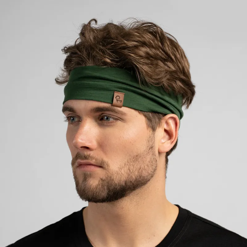 A close-up studio portrait of a man looking to the left against a plain light grey background. He has wavy brown hair held back by a dark green headband and is wearing a black shirt.