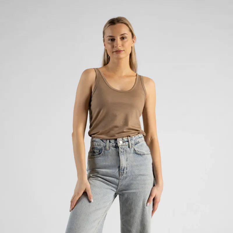 A studio portrait of a blonde woman standing and looking directly at the camera with a subtle smile. She is wearing a beige sleeveless scoop-neck top and light wash denim jeans, with one hand casually resting in her front pocket against a plain light grey background.